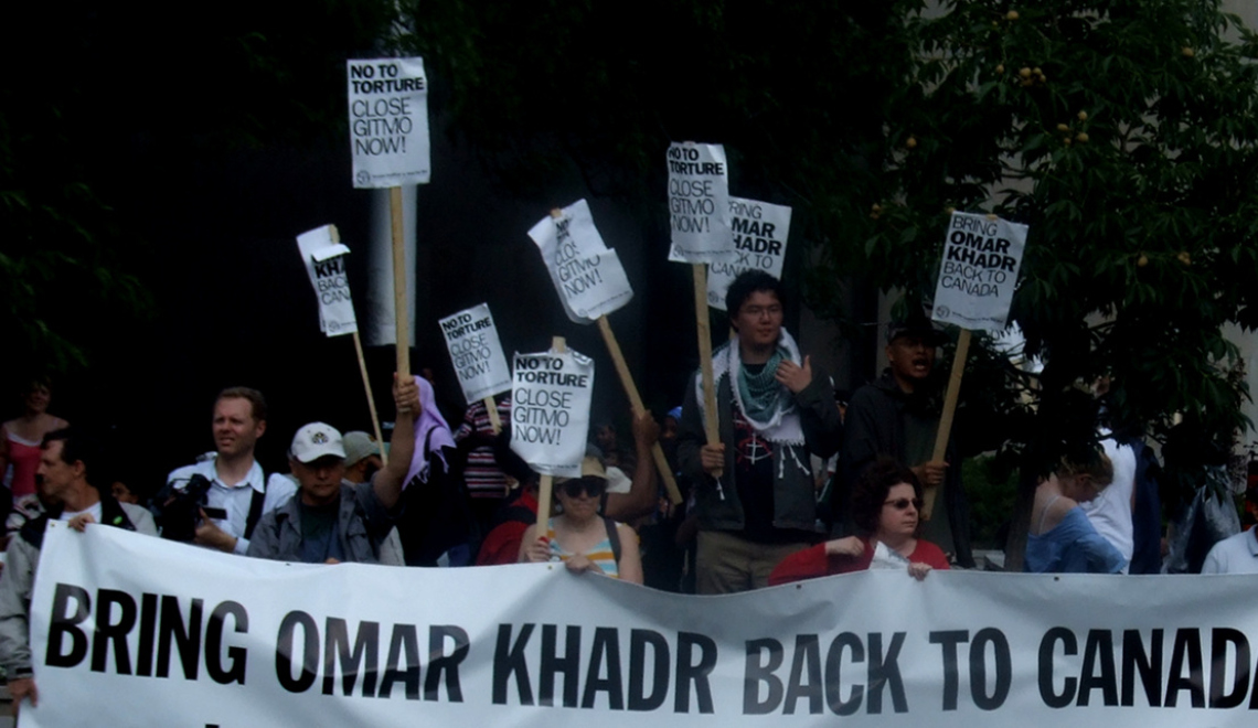 Omar Khadr Banner - Joshua Sherurcij - Wikimedia Abbildung von Demonstranten mit Banner